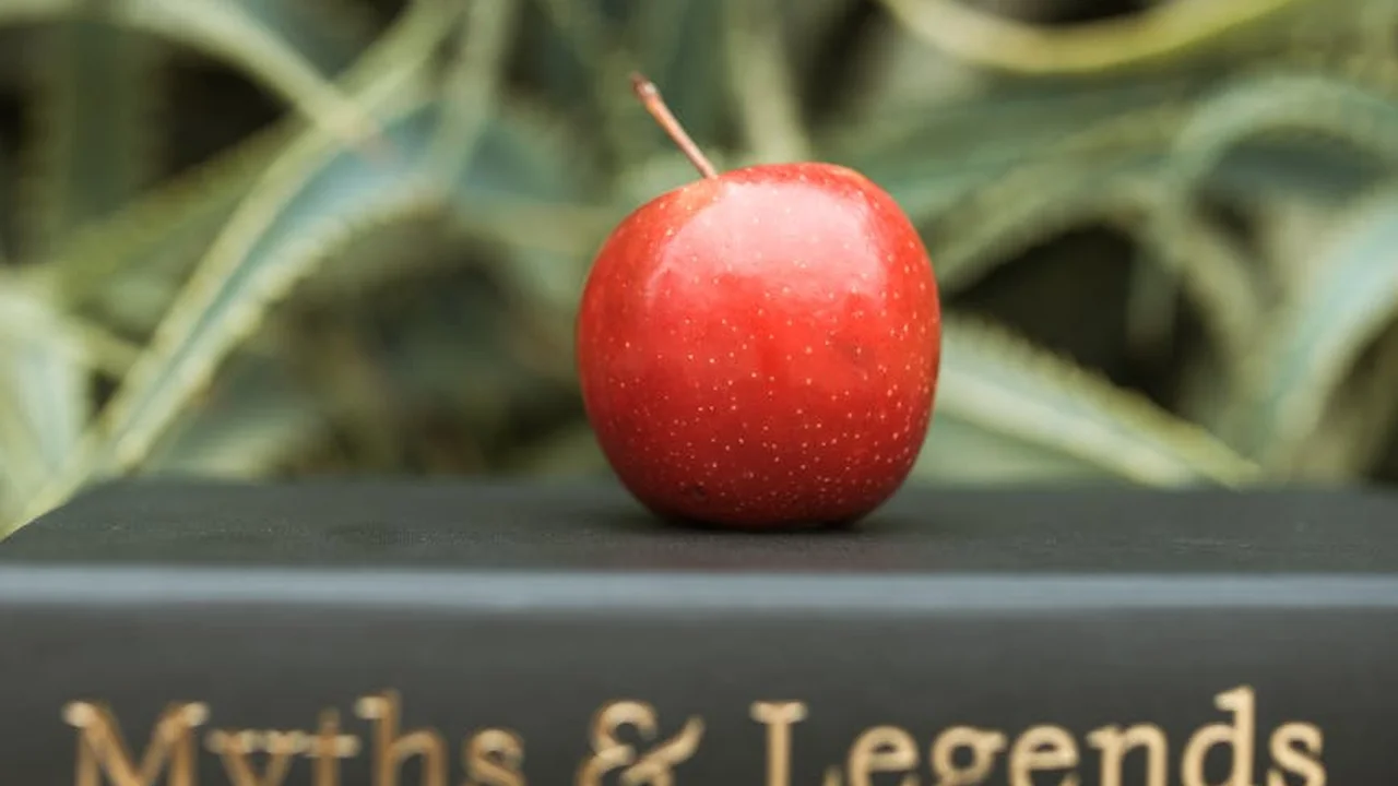 A bright red apple rests on top of a dark book spine with gold lettering, with blurred green foliage in the background.
