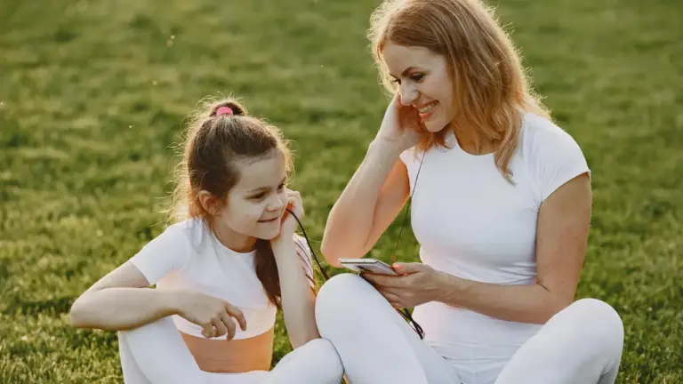 A woman and a young girl sit on a grassy field outdoors, looking at a smartphone together as they listen to an audiobook.