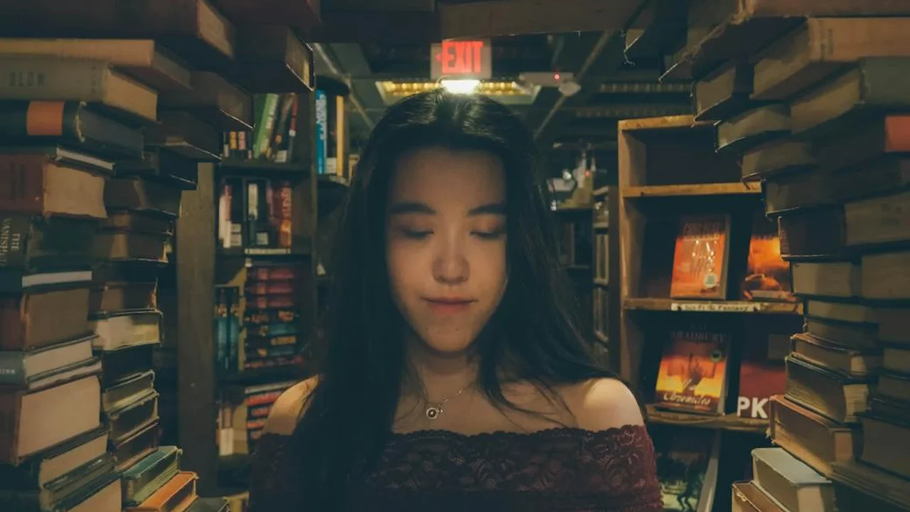 A young woman stands in a cozy bookstore aisle, surrounded by towering shelves of books, looking down with a soft expression.