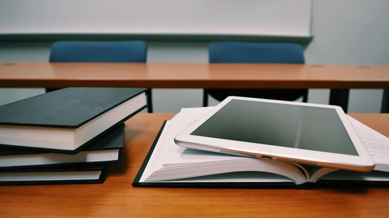 A classroom desk with an open book, a tablet resting on the pages, and several stacked notebooks nearby.