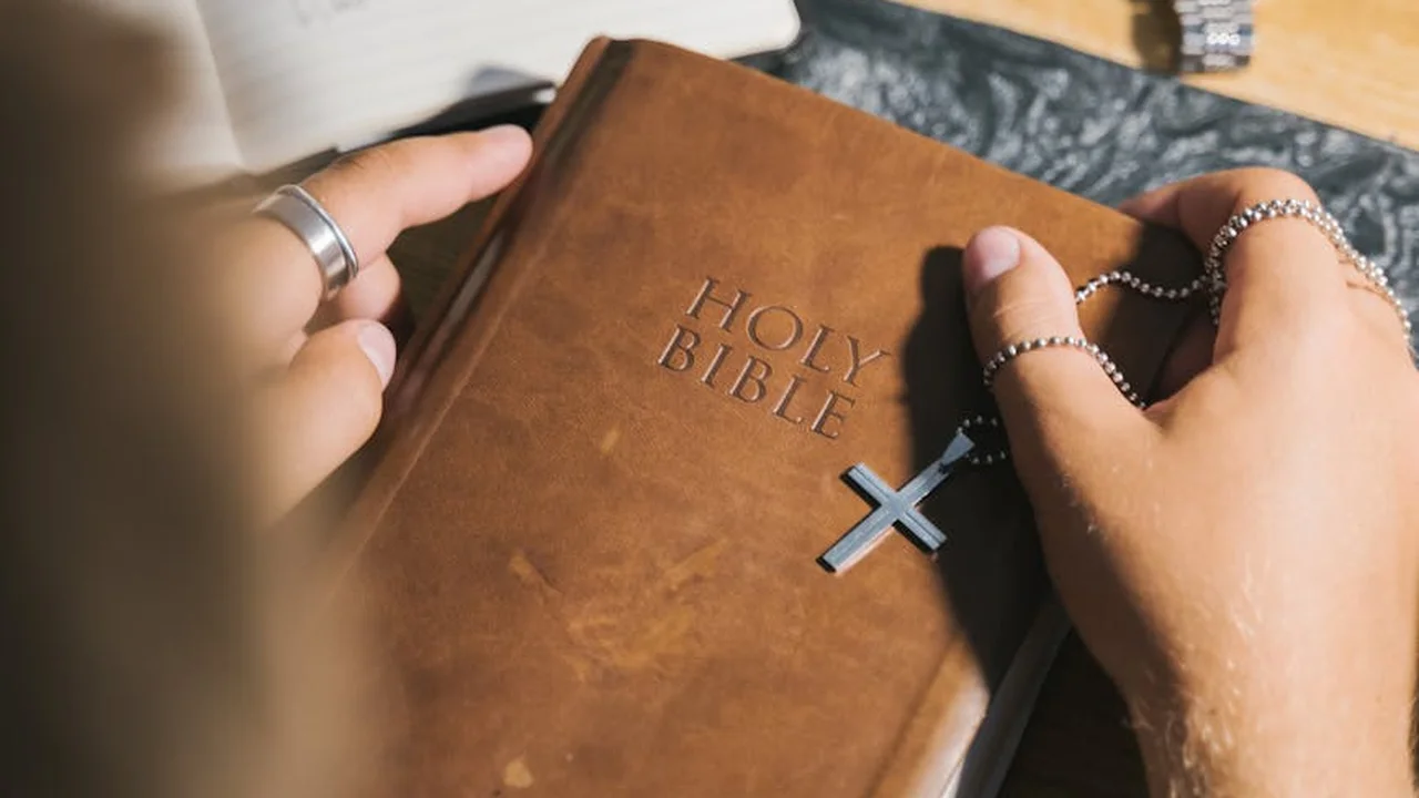 Close-up of a person’s hands holding a brown Holy Bible with a silver cross necklace resting on the cover.