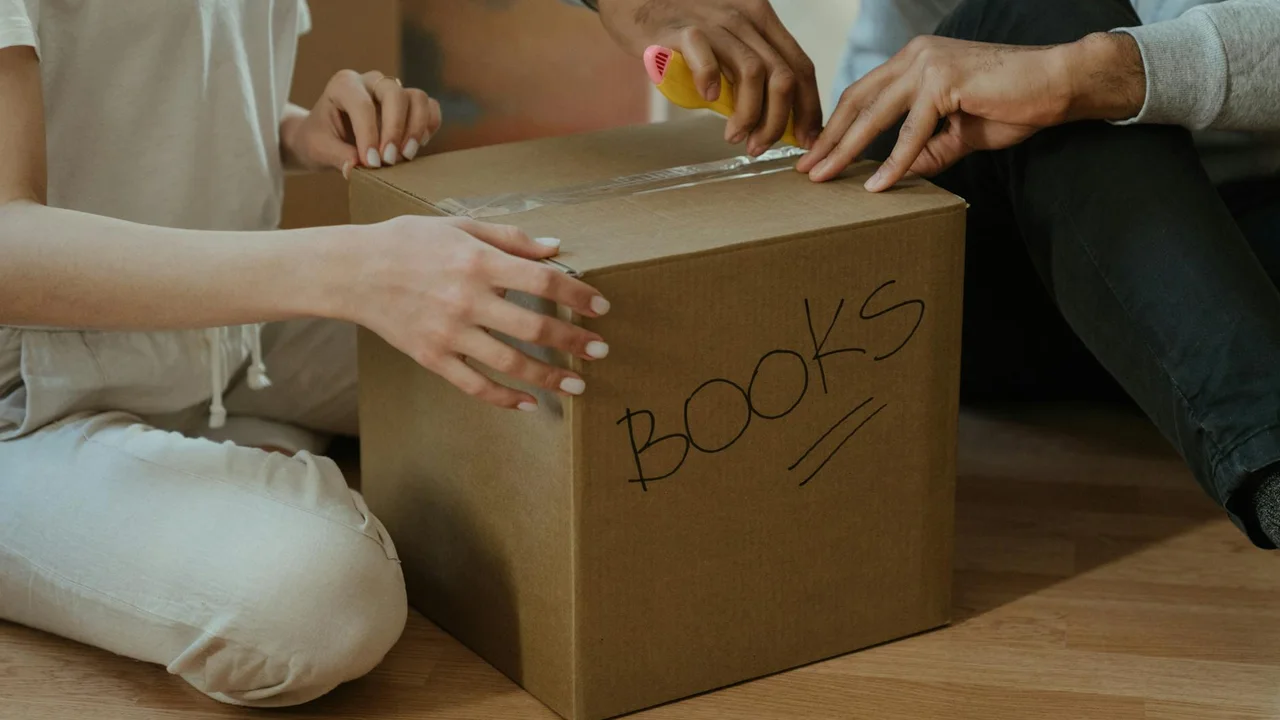 Two people sit on a wooden floor and open a cardboard box labeled 'BOOKS'.