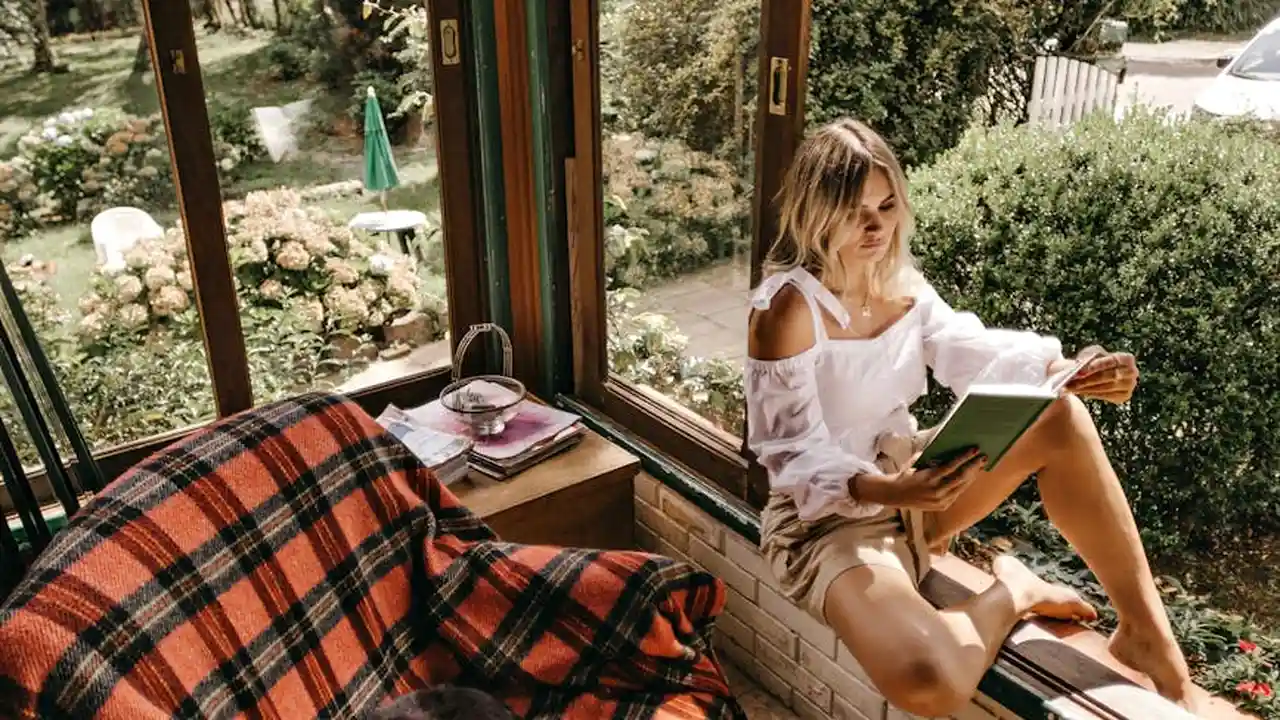 Woman wearing a white top and beige shorts sits by a sunlit window reading a book, with a plaid blanket on a nearby chair.