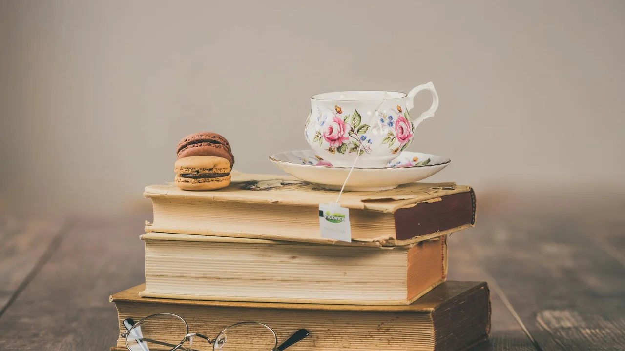 A stack of vintage hardcover books with a floral teacup on top and a macaron to the side on a wooden table.