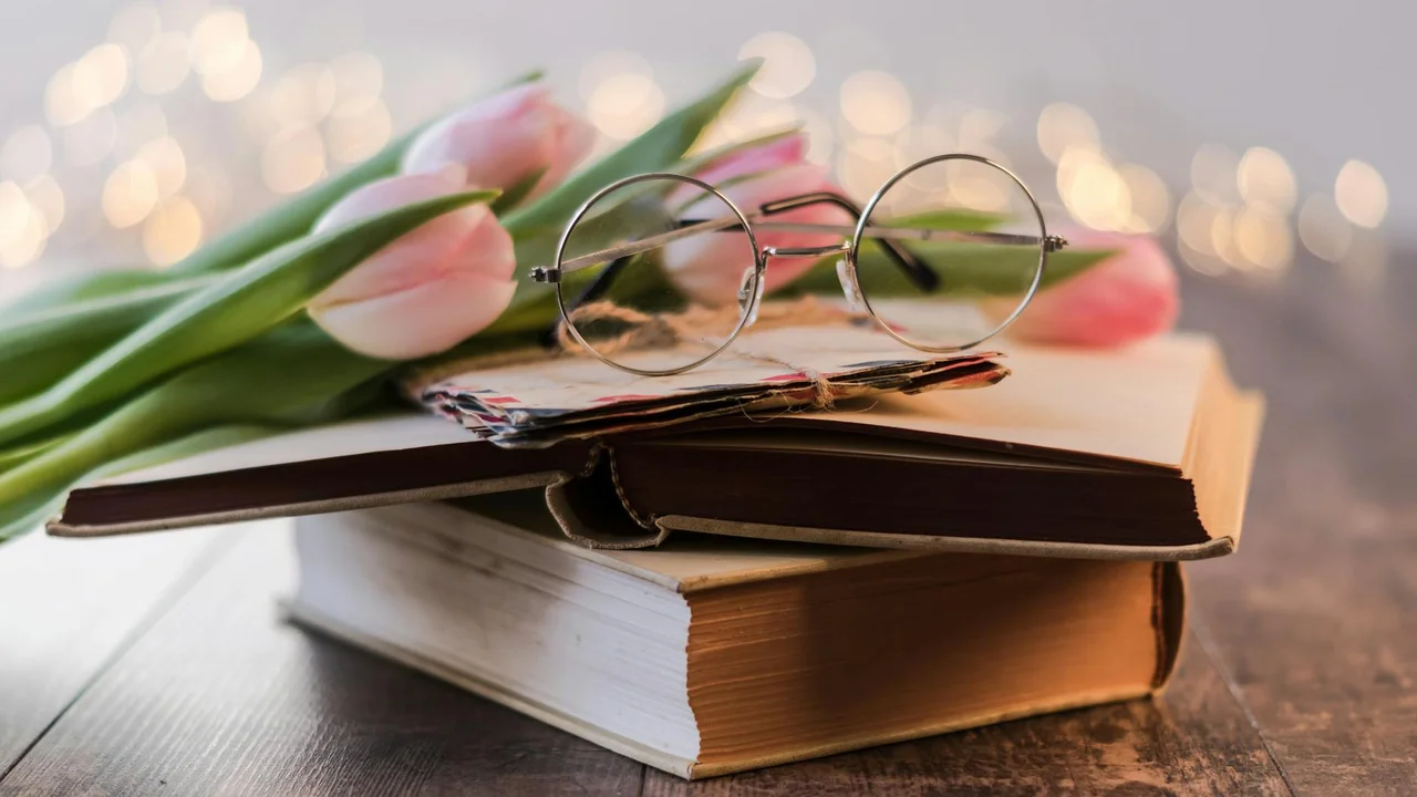 A stack of books with a pair of round wire-rimmed glasses resting on top, pink tulips to the side, and warm bokeh lights in the background.