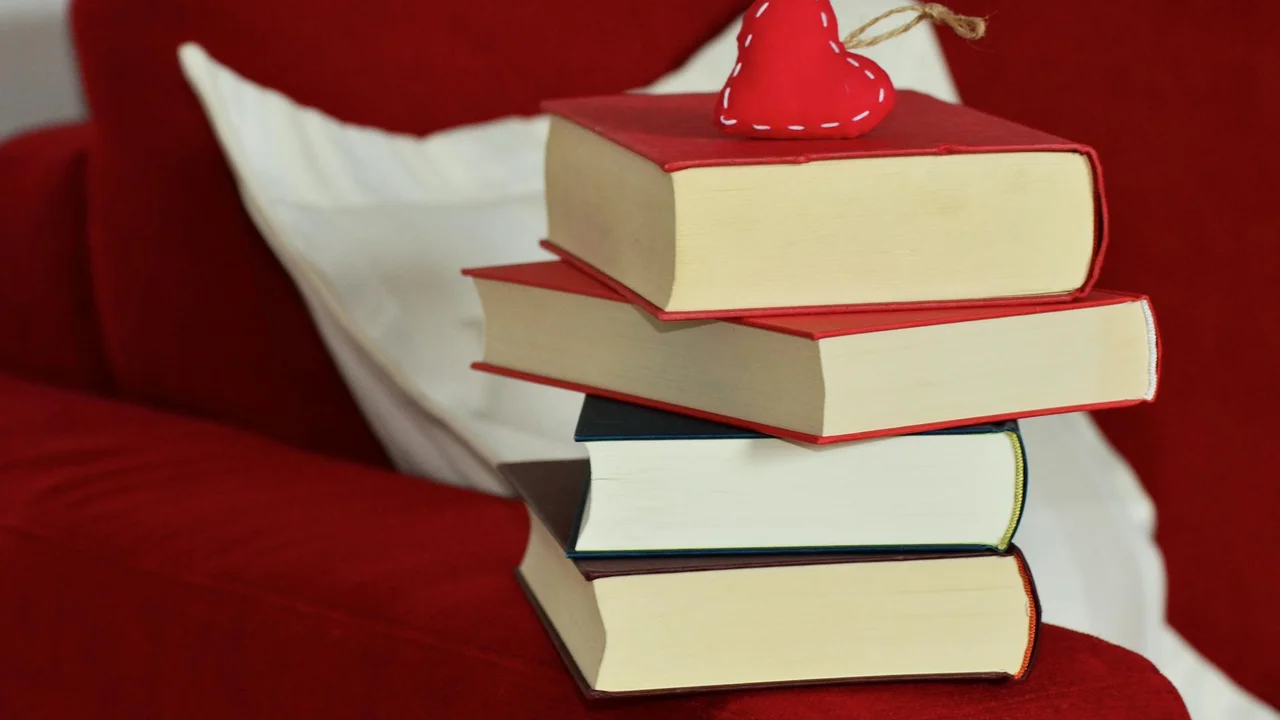 Stack of hardcover books on a red couch with a heart-shaped ornament on top