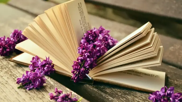 An open book with its pages fanned out on a wooden surface, adorned with purple flowers