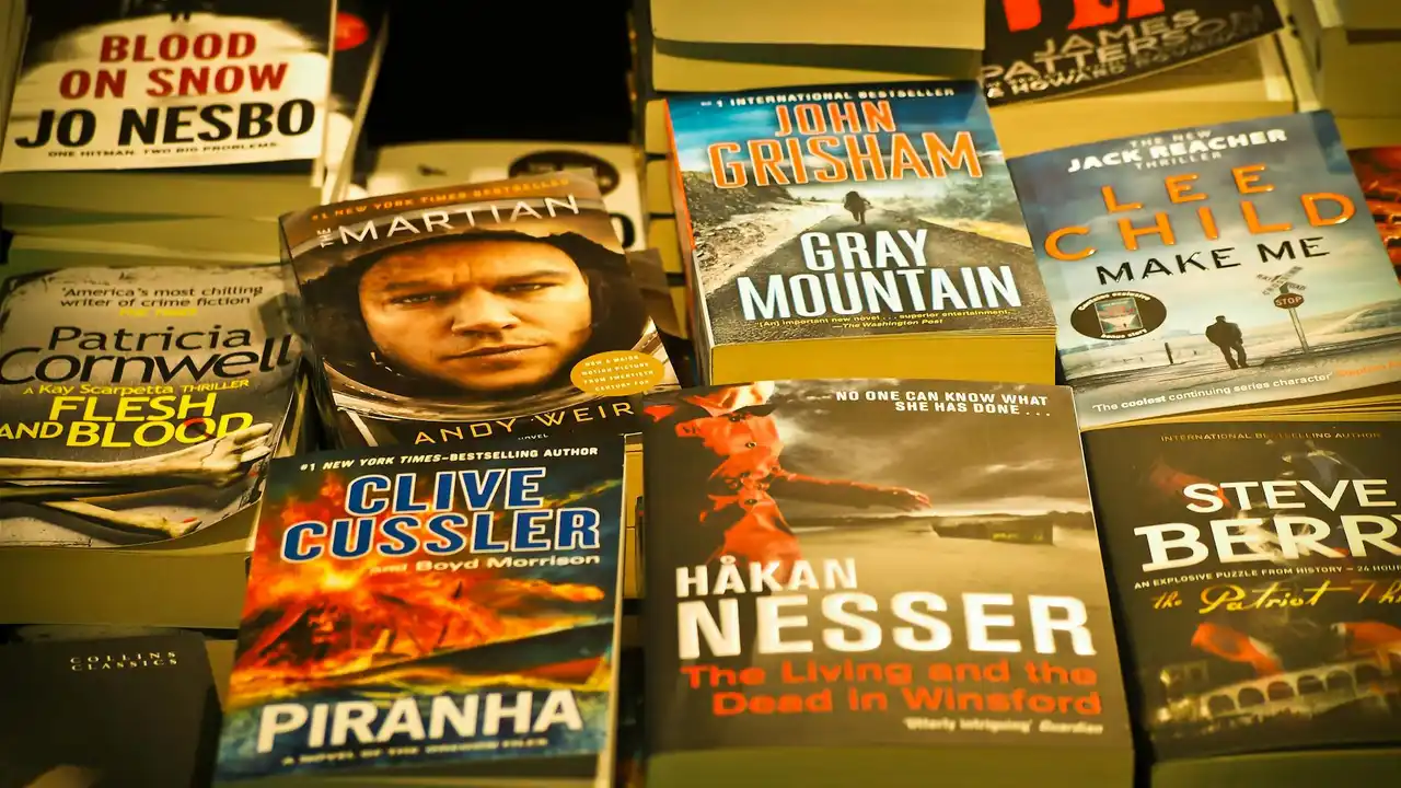 A close-up collage of thriller and crime book covers on a bookshelf, featuring titles such as Gray Mountain, Piranha, Make Me, Blood on Snow, and The Living and the Dead in Winter.