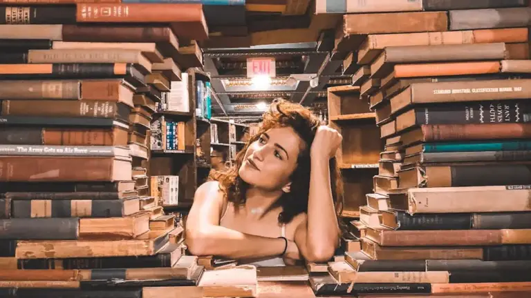 A woman rests her head on her arm in a cozy passage formed by stacked book spines, surrounded by shelves of books.