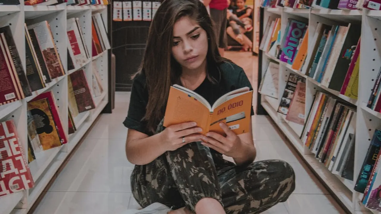 Young woman sitting cross-legged in a bookstore aisle, reading an orange paperback