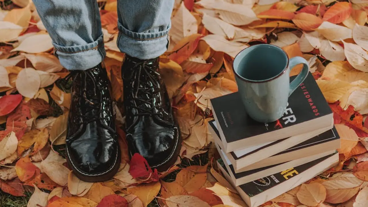 Close-up of black, shiny ankle boots standing on a bed of orange and yellow autumn leaves, with a small stack of books and a blue mug resting on top.