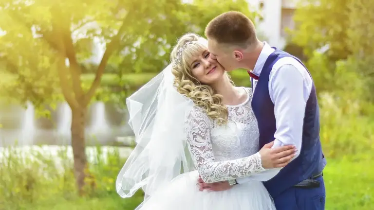 A bride in a lace wedding dress and veil embraces a groom wearing a vest and tie in a sunlit park.