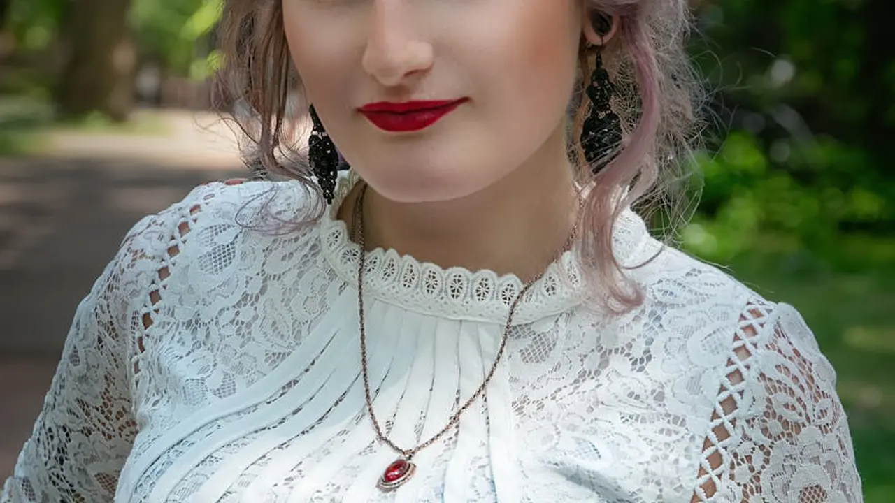 Portrait of a woman with light skin wearing a white lace high-neck blouse and bold red lipstick, outdoors with greenery in the background.