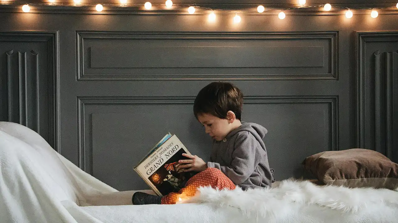 A young child sits up in bed, reading a book under warm string lights, with a fluffy blanket and pillows beside them.