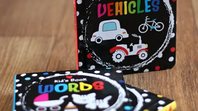Two small black-covered children's books with colorful word titles 'Words' and 'Vehicles' on a wooden surface, featuring playful illustrations of a tractor and a bicycle.