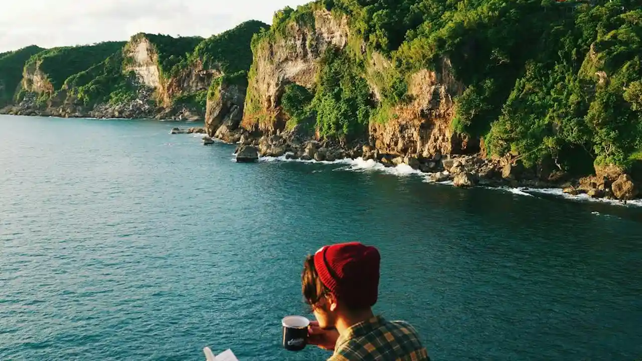 Person in a red beanie standing on a rocky shoreline, looking toward a calm blue sea with green cliffs in the background.