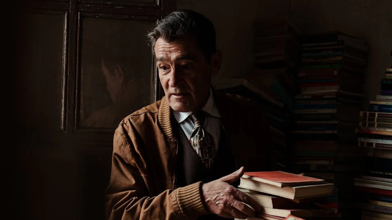 A man in a brown cardigan stands in a dimly lit library, holding a stack of books.