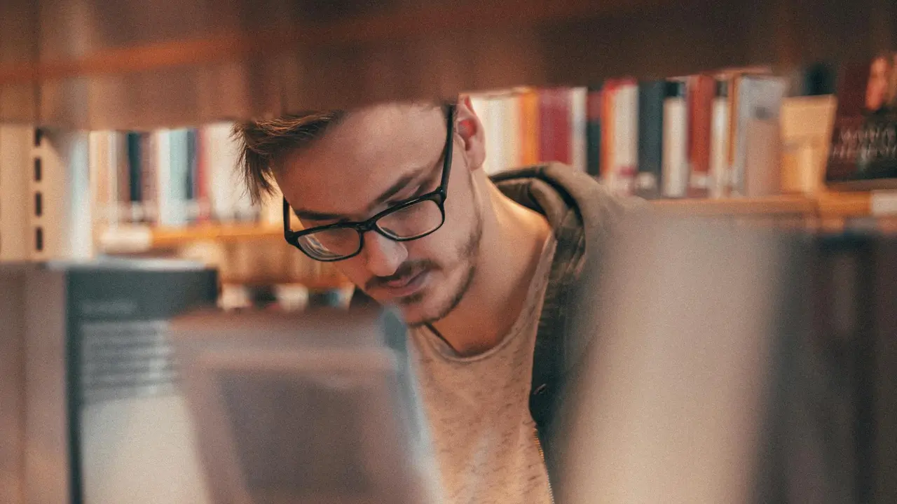 Young adult with glasses in a library, looking down at a device on a table, with bookshelves blurred in the background.