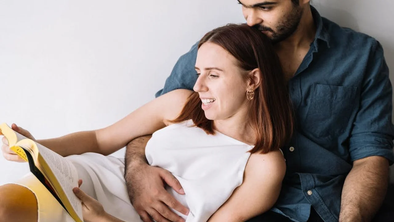 A couple cuddles on a couch while the woman reads a book, the man embracing her from behind
