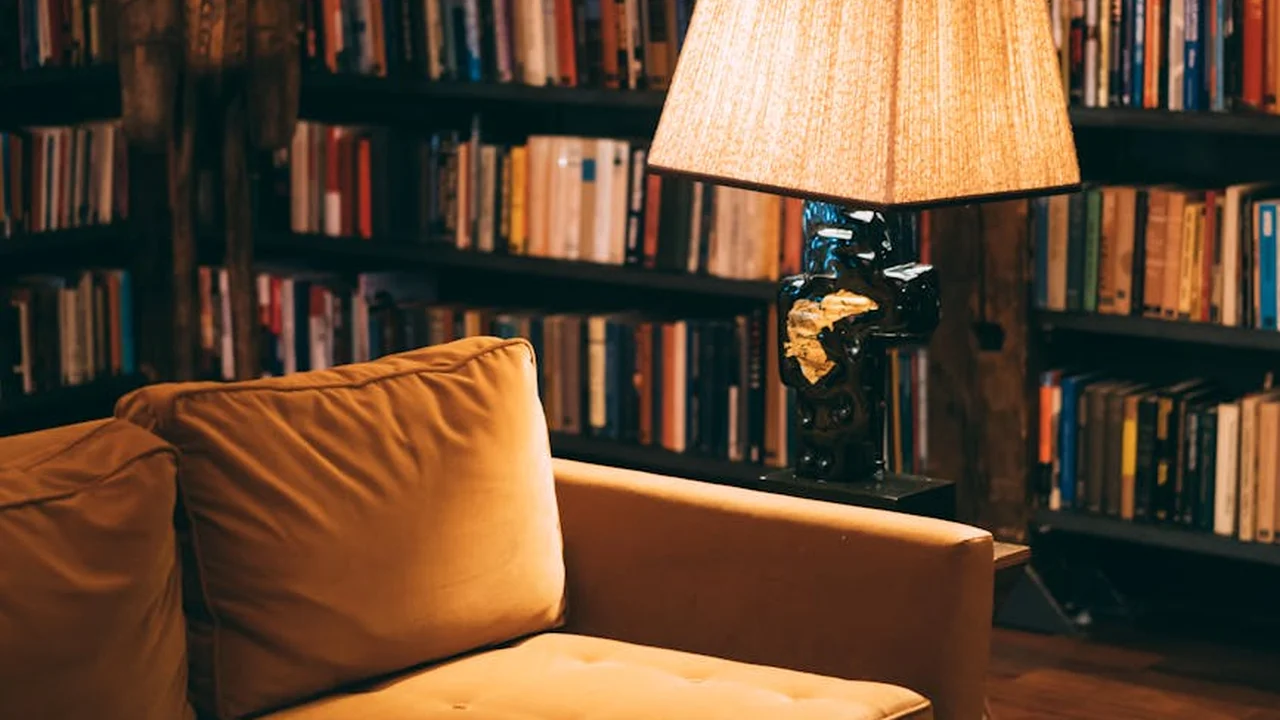 A cozy library scene with a warm-lit lamp on a side table beside a mustard-yellow sofa, surrounded by tall bookshelves filled with books.