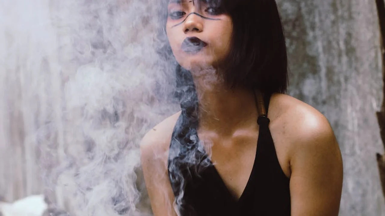 Portrait of a woman with short black hair wearing a black halter top, exhaling smoke in a moody, textured setting.