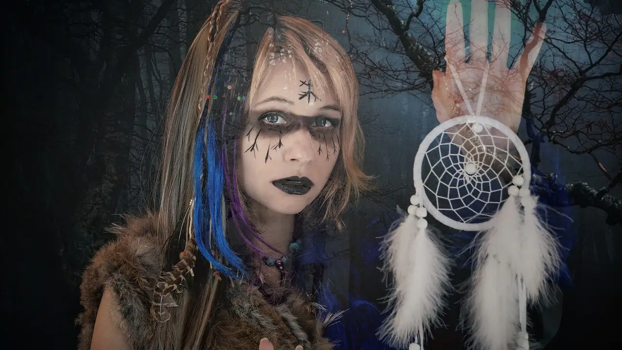 Portrait of a young witch with pale makeup and black lips, rune-like markings on her face, blue and purple hair strands, wearing fur and beads, holding a white dreamcatcher against a dark, moonlit forest.