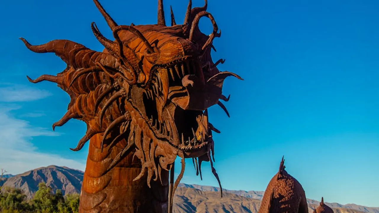 Rust-colored dragon sculpture with an open mouth and multiple horns, set against a bright blue sky and distant mountains.