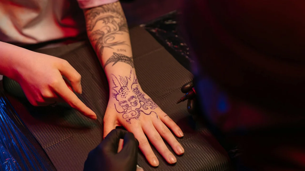 Close-up of a tattooed forearm and hand resting on a dark surface, featuring a dragon tattoo and intricate linework.