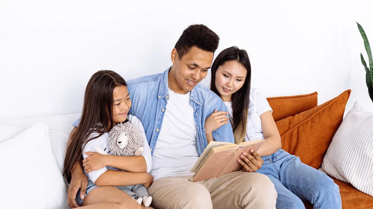 A family sits together on a white sofa, reading a book. The father and mother lean in with their daughter, who holds a stuffed animal.