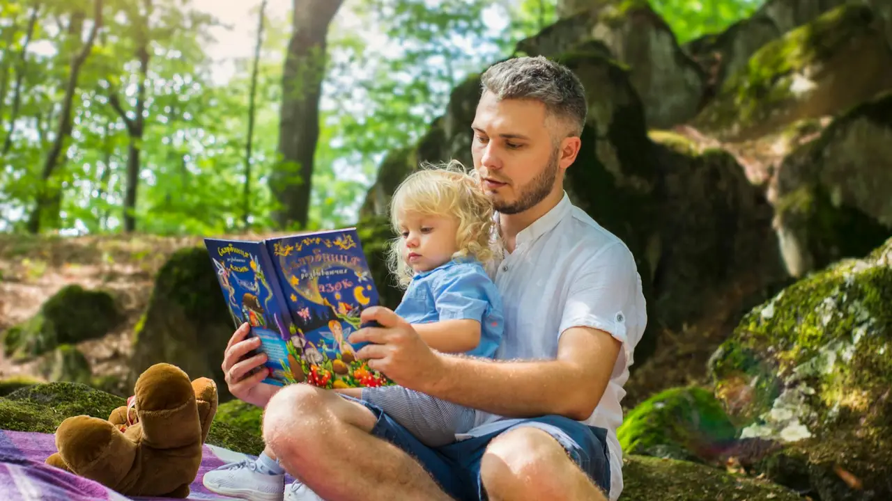 A man sitting on the grass in a park reads a colorful children's book aloud to a young girl on his lap.