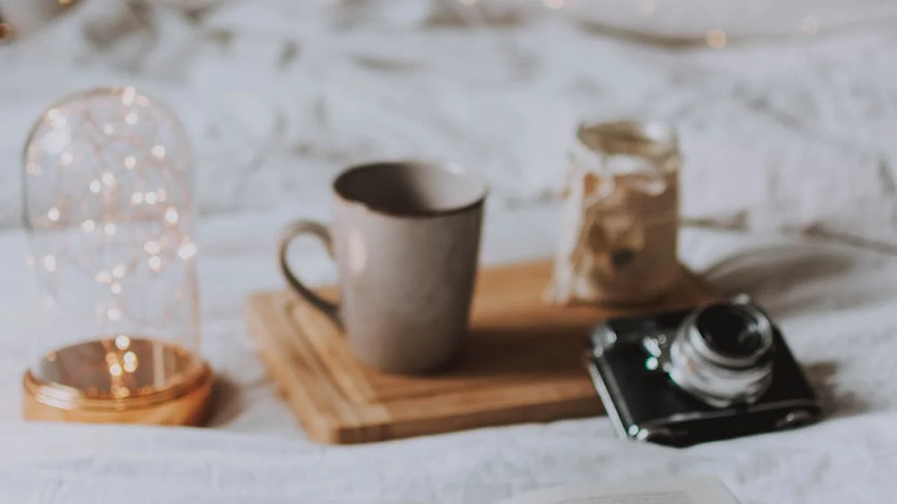 Minimalist still life with a camera, mug, wooden tray, and twinkling fairy lights under a glass cloche on a light surface.