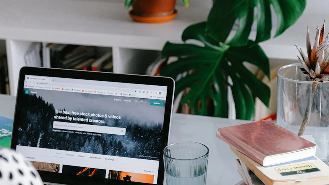 A laptop on a white desk with a plant, books, and a glass of water, conveying a cozy home workspace.