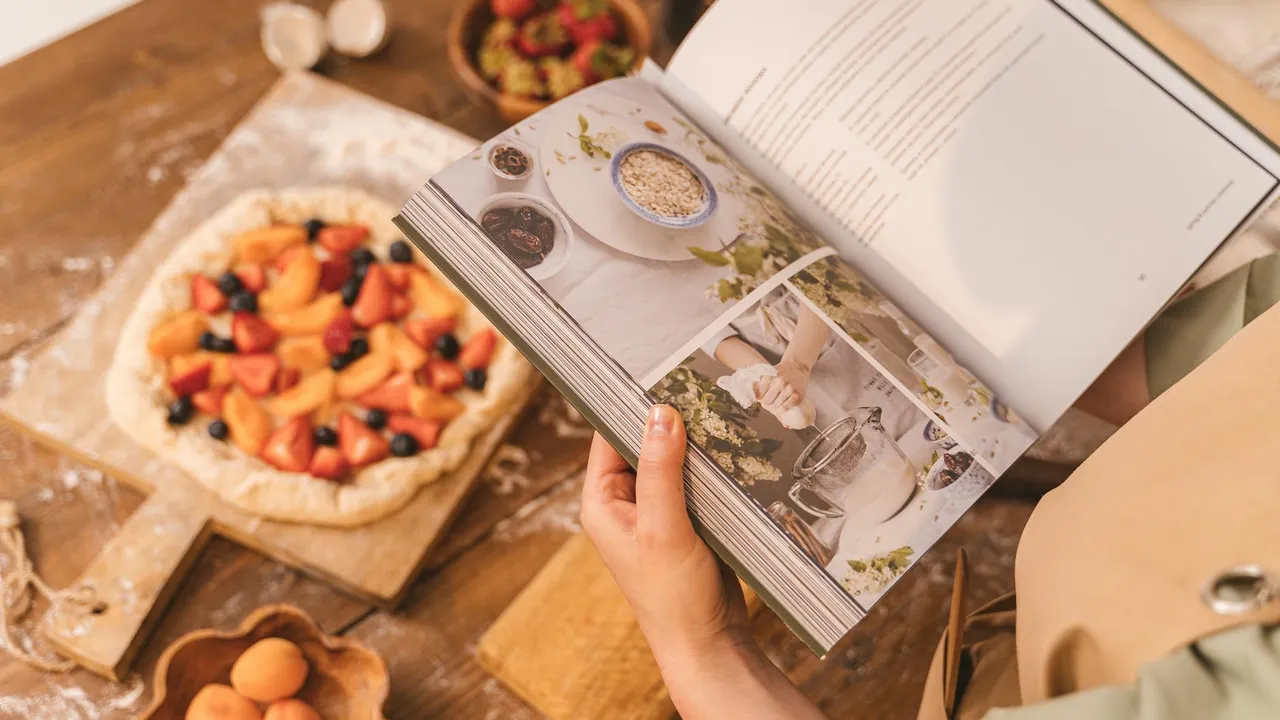 Person flipping through a cookbook while a homemade pizza rests on a wooden table.