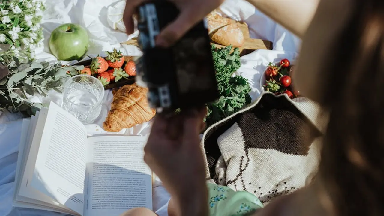 A person taking a photo of a picnic spread on a blanket, featuring an open book, croissants, apples, herbs, and other food items.