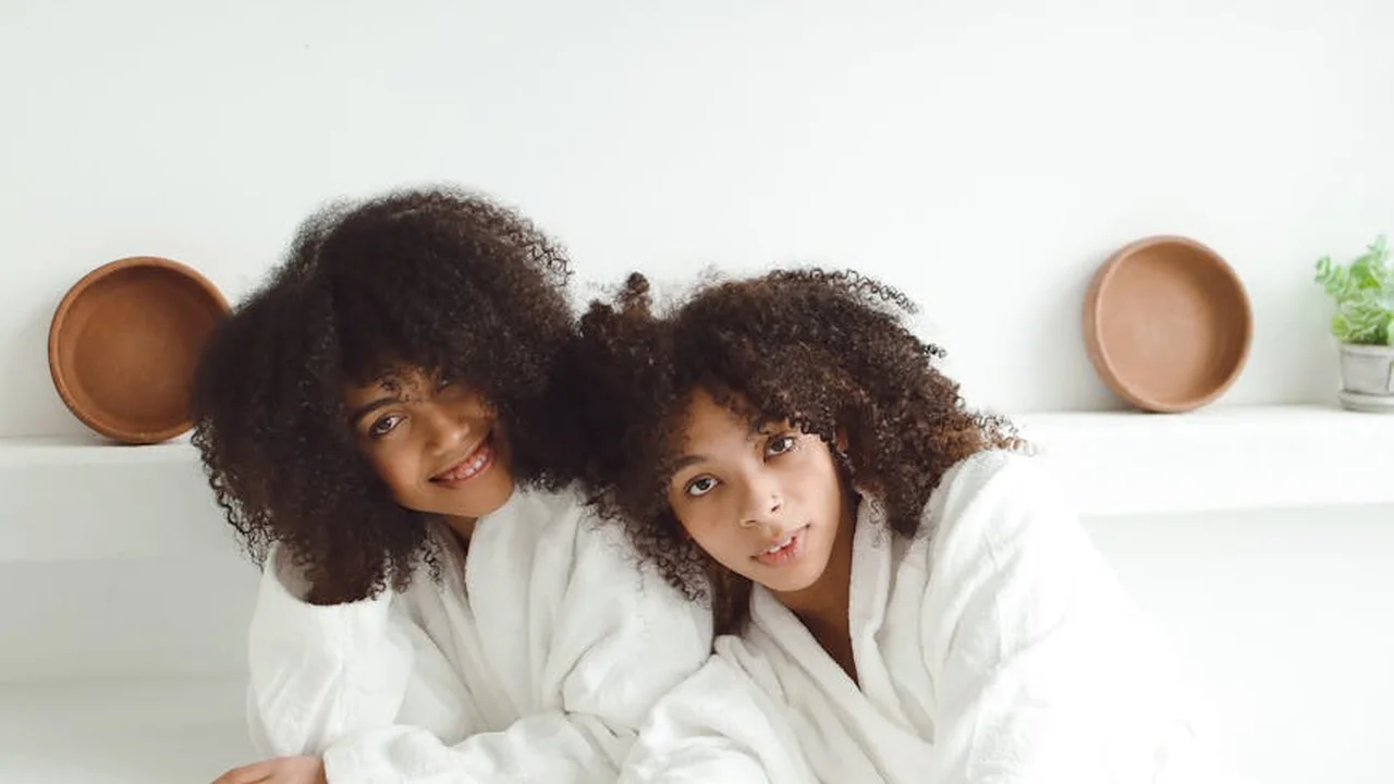 Two young sisters with curly hair, wearing white robes, posing together indoors and smiling.