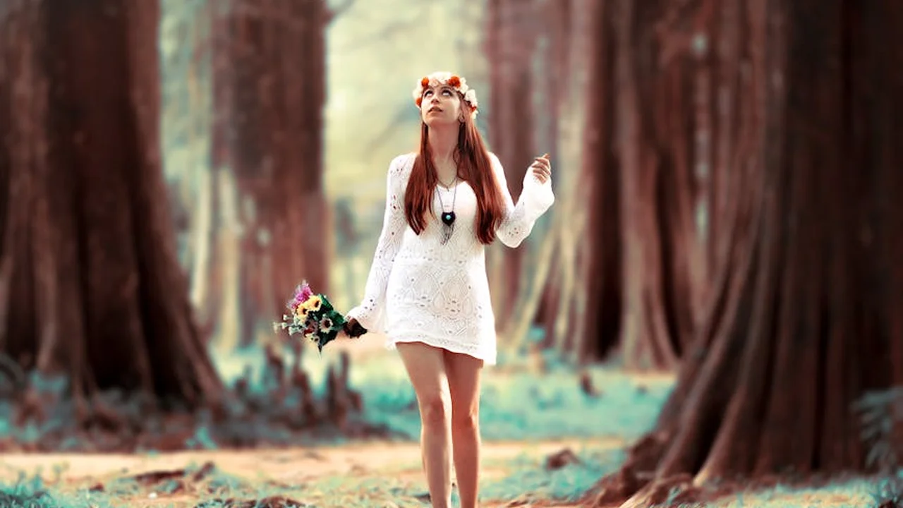 A woman wearing a white lace dress and a flower crown stands in a sunlit forest, holding a small bouquet as tall trees surround her.