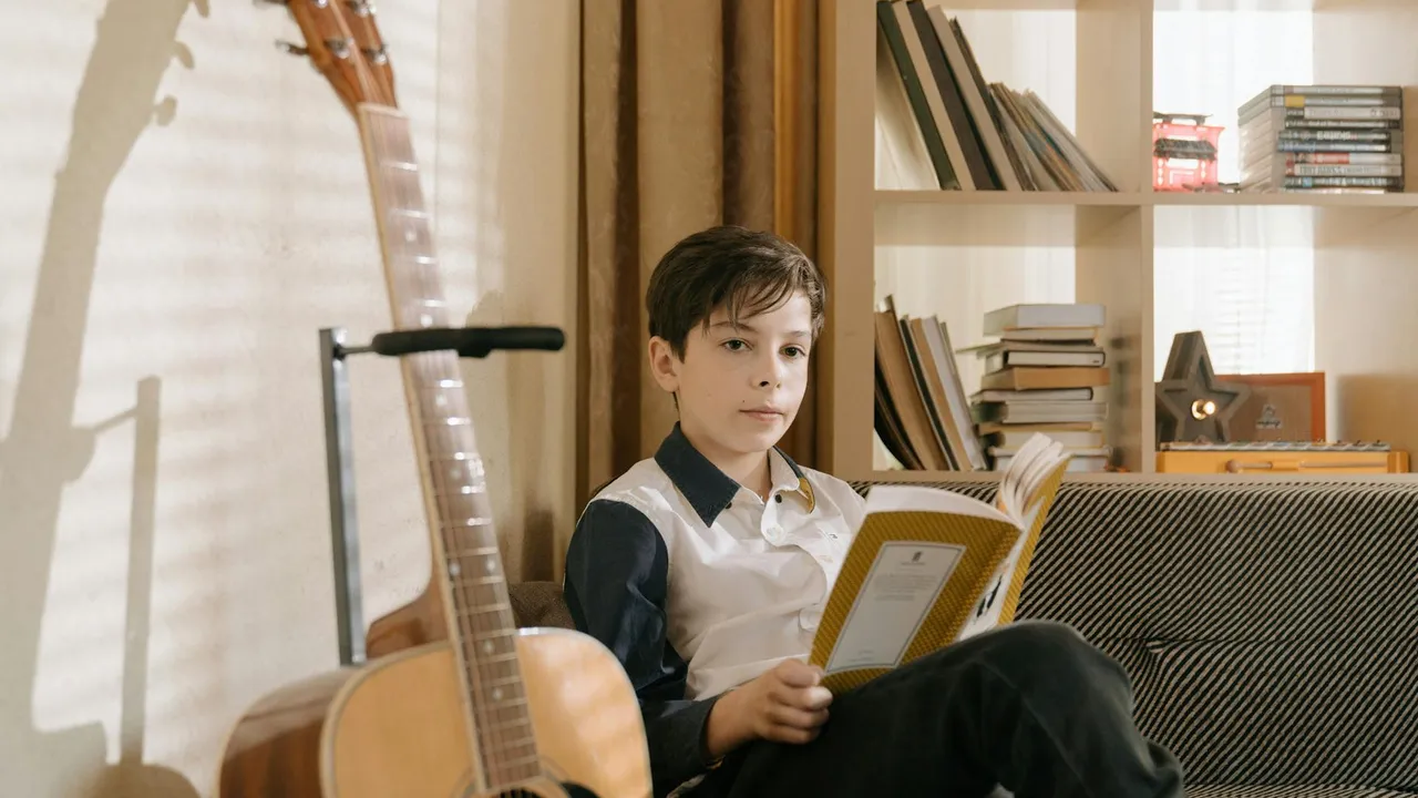 A child sits on a sofa reading a book, with a guitar leaning against the wall and a bookshelf in the background.