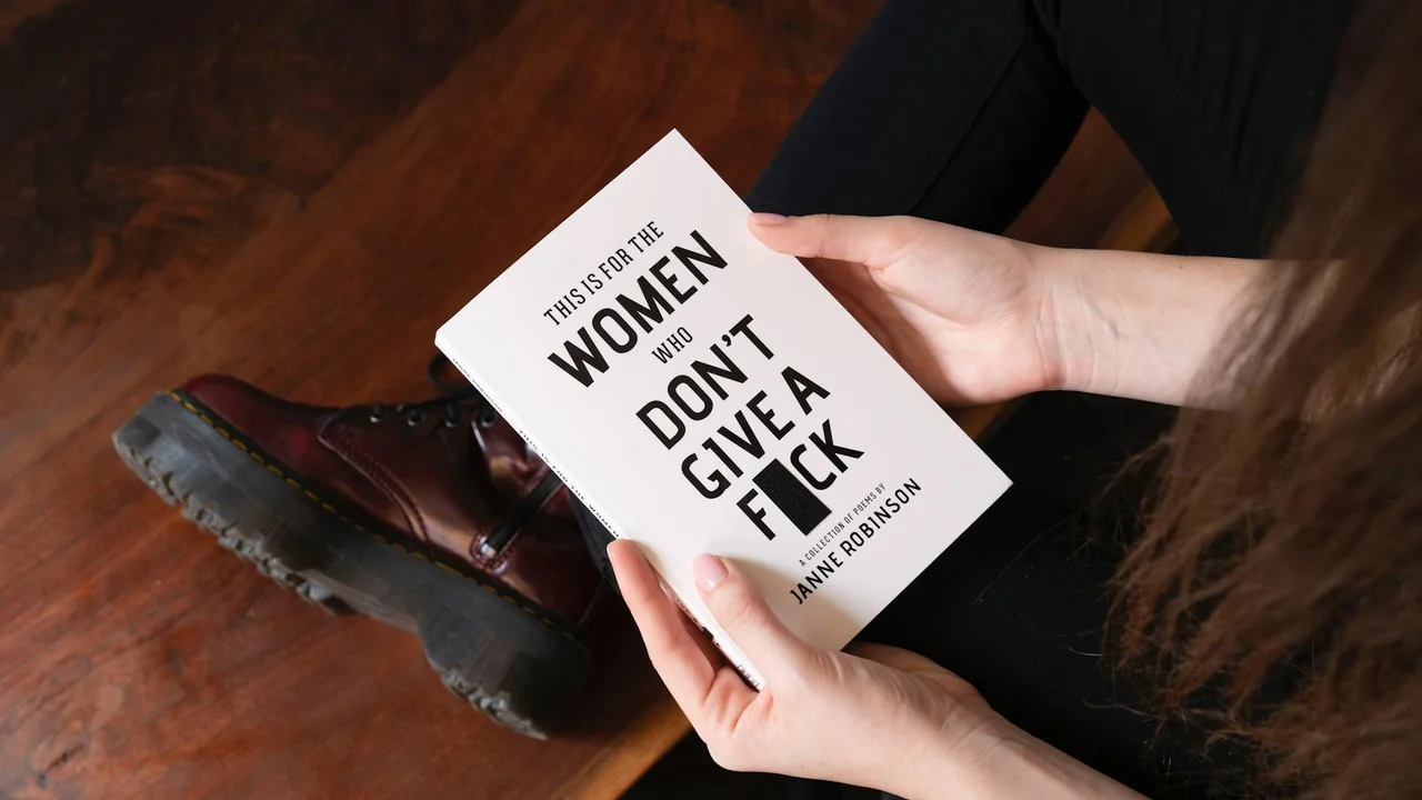 Person sitting on a wooden floor, holding a white paperback with bold black text on the cover.