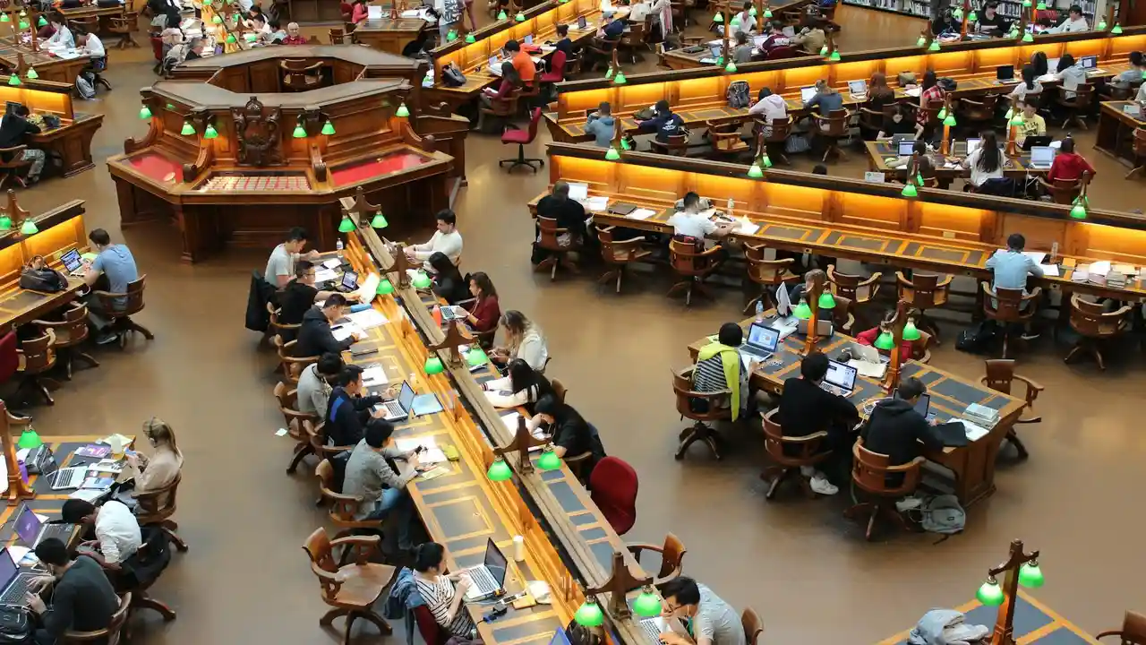 Busy library reading area with patrons seated at long, curved tables, reading books and using laptops.