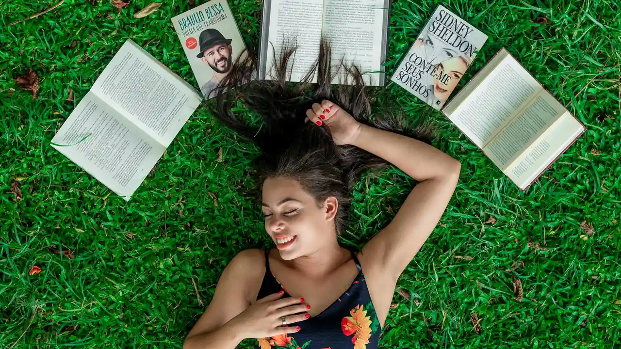 Young girl lying on vibrant green grass, surrounded by open books, smiling with eyes closed.