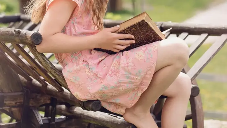 Young girl in a pink floral dress sits on a park bench, reading a hardcover book.
