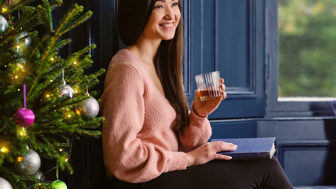 A smiling woman with long dark hair sits by a window next to a decorated Christmas tree, holding a glass in one hand and a book in the other, in a warm, cozy holiday room.