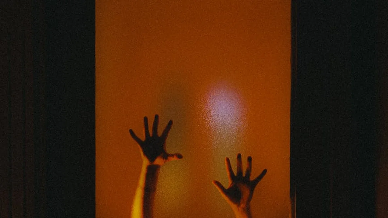Two hands pressed against a glass pane in a dark, orange-lit corridor, creating a jump-scare atmosphere.
