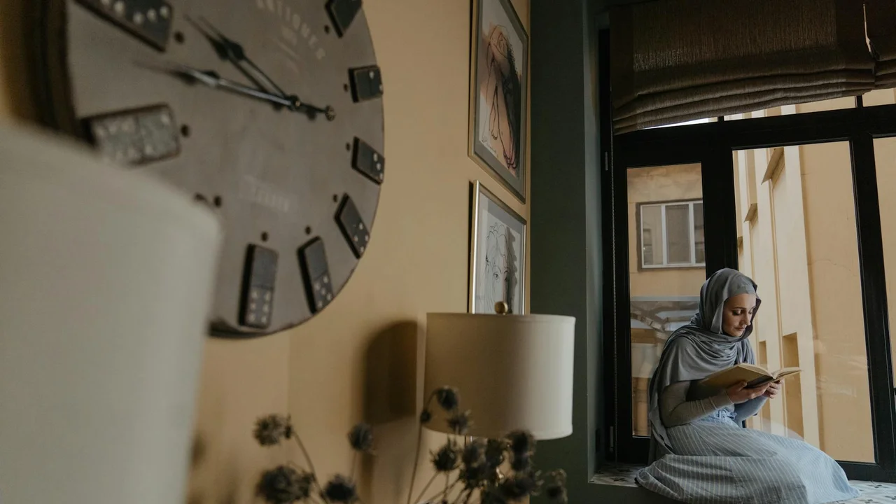 Young woman wearing a light gray hijab sits by a window reading a book in a cozy interior.