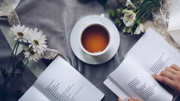Top-down view of a reading setup: a cup of tea on a saucer, white daisies, lace, and two open books with a person turning pages