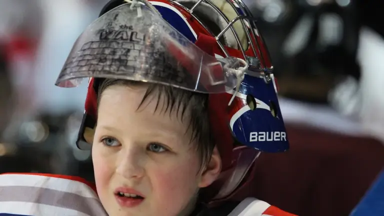 A young hockey goalie wearing a protective helmet and pads, focused on the action on the ice.