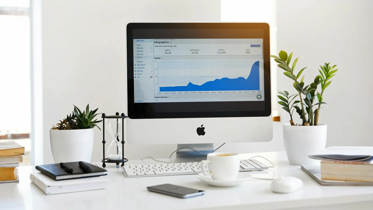 Desk setup with an iMac displaying graphs, surrounded by plants, books, a notebook, and a coffee cup.