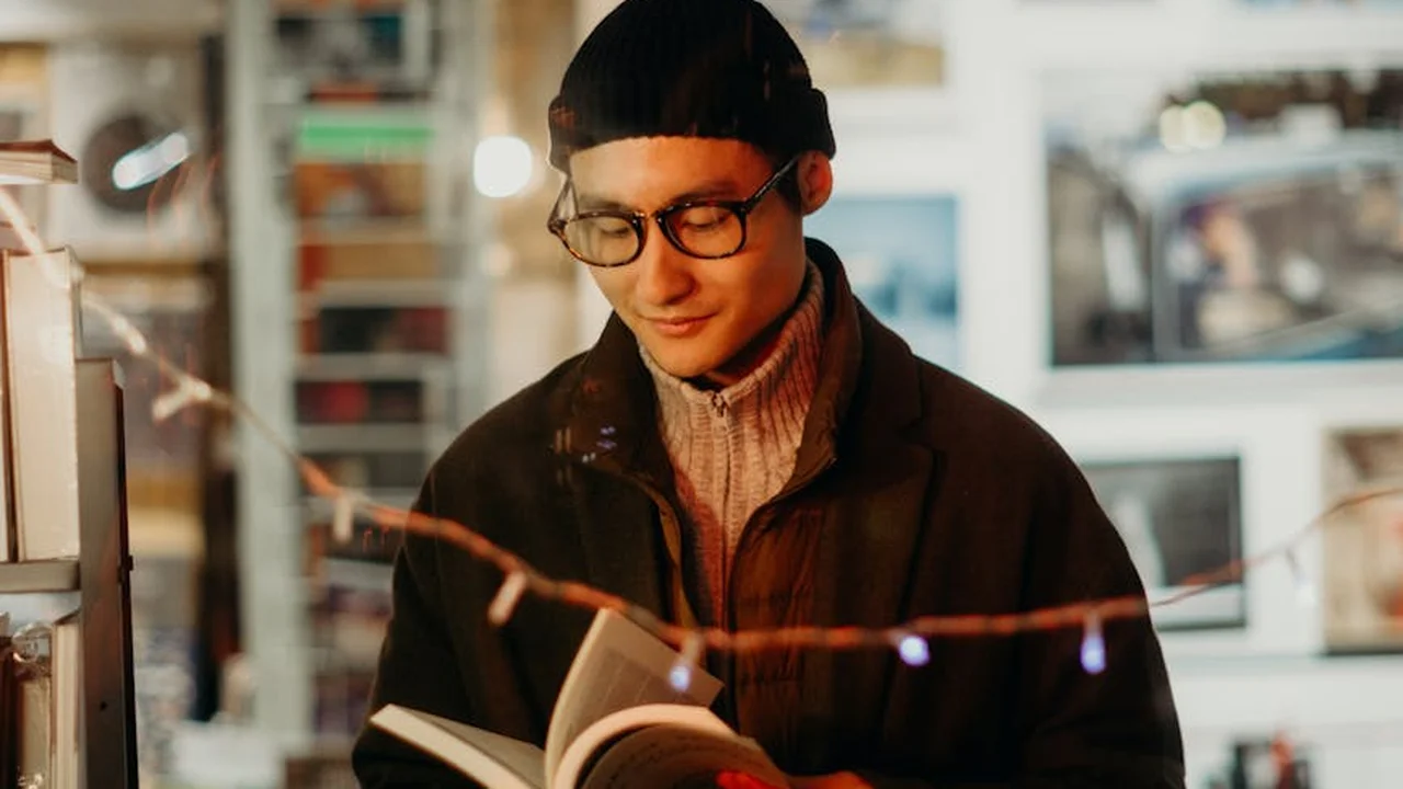 A young man wearing a beanie and glasses reads a book in a cozy bookstore, with string lights and shelves in the background.