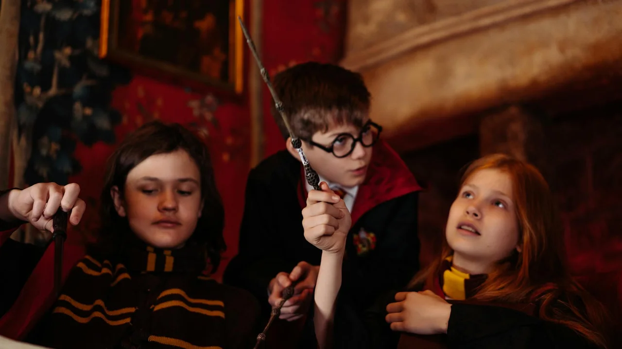 Three children in dark robes gather around, with one holding a wand upright. They are in a dimly lit room with warm, medieval-inspired decor.