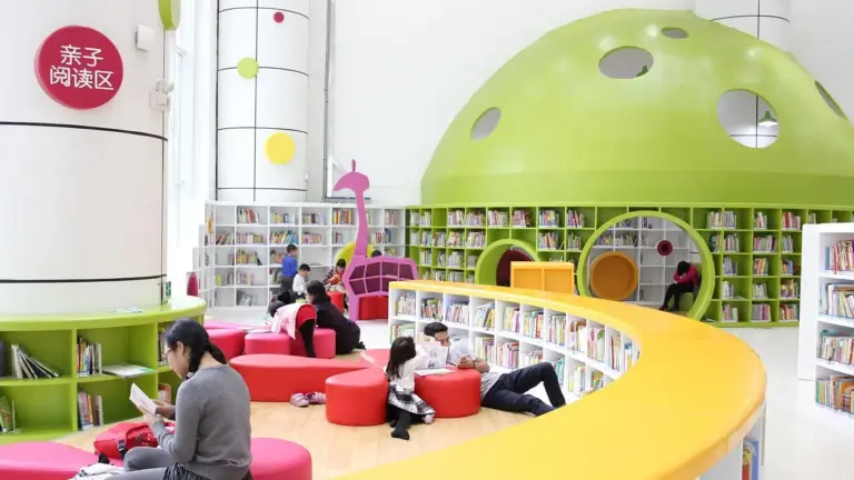 Children sit and read in a bright, modern children's library with curved yellow seating and a green dome ceiling.